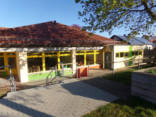Auf dem Bild ist die Rückseite des "Kindergarten Meerhof" zu sehen. Das Foto zeigt einen gepflasterten Weg der in einer Treppe mündet und eine Rutsche.